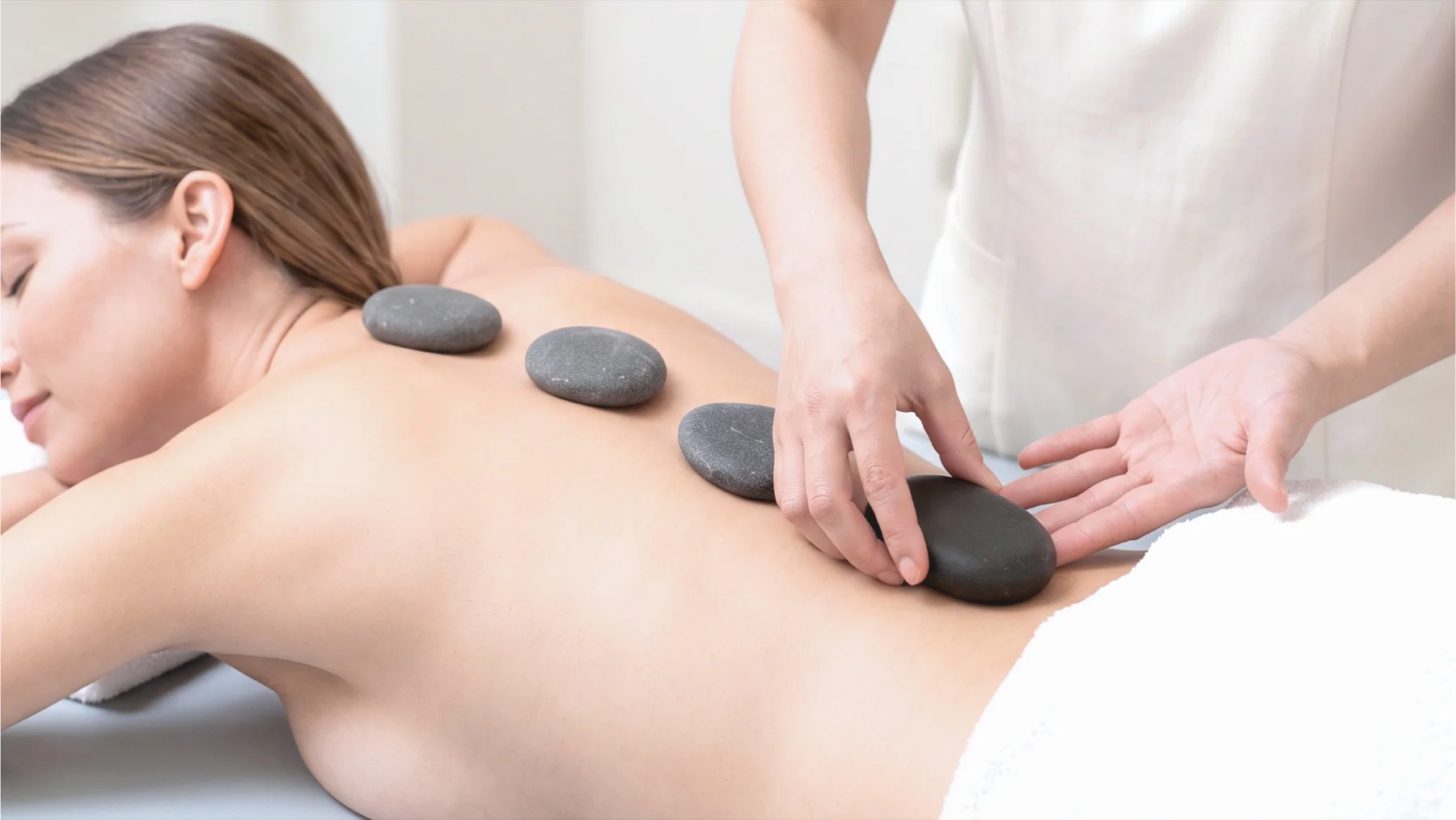 Smooth warm stones arranged neatly beside a therapist performing a professional massage, in a serene but realistic home setting. Avoid visible steam or intense red skin.